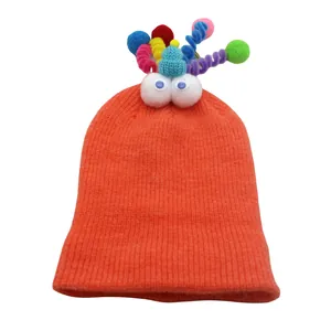Detail of orange knitted hat featuring plush eyes and colorful pom-pom antennas