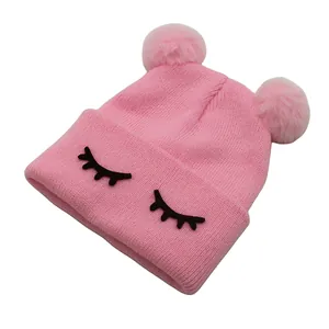 Detail van roze gebreide babymuts met dubbele pompons en een geborduurd oogjespatroon erop