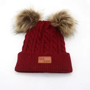 Close-up of wine red cable knit beanie featuring fluffy double pom-poms and leather logo