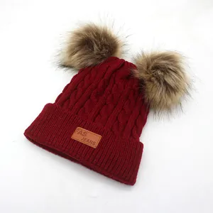 Detail of burgundy cable knit kids hat with dual faux fur pom-poms and leather logo patch