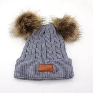 Grey knit winter hat for children featuring double faux fur pom-poms and leather logo patch