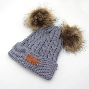 Detailed close-up of grey cable-knit kids' hat with dual pom-poms and brown logo patch