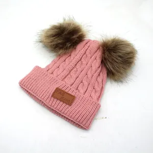 Close-up of pink knit hat with dual brown pom-poms and leather logo patch detail