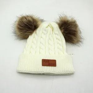 Detail of cable knit kids winter hat with two faux fur pom poms and leather logo patch