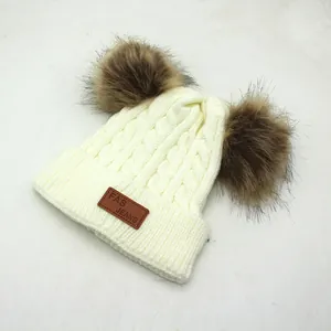 Detail of cable knit beanie with dual brown faux fur pom-poms and leather logo patch