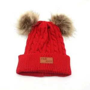 Red cable knit winter hat for children featuring dual fluffy faux fur pom poms and leather patch