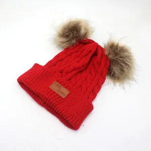 Close-up of red cable knit kids beanie with double faux fur pom-poms and leather patch