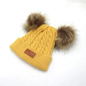 Detail of yellow knit hat featuring double faux fur pom-poms and a leather brand patch