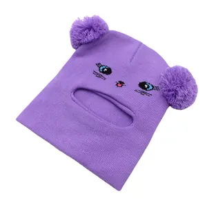 Dettaglio di passamontagna in maglia viola con viso di gatto e grandi pompon morbidi