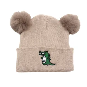 Beige knit winter hat for babies featuring crocodile embroidery and fluffy pom-poms