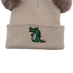 Detail of khaki knit baby hat with dual brown fur pom-poms and green crocodile embroidery