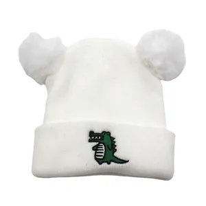 White knit baby beanie with large faux fur pom-poms and green crocodile embroidery