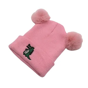Detail of pink knitted baby hat with soft faux fur pom-poms and green dinosaur embroidery
