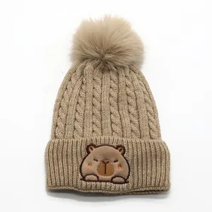 Close-up of khaki knit beanie featuring a fluffy pompom and cute capybara patch detail