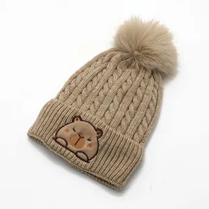 Detail of knitted winter hat with capybara embroidery and large faux fur pom-pom