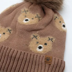 Close-up of coffee brown knit hat with fluffy pom-poms and cute bear embroidery details