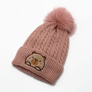 Detail of pink knitted beanie featuring cute capybara embroidery and fluffy pom pom