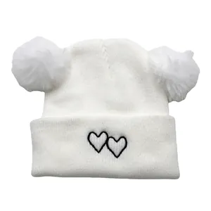 Close-up of white knit baby hat featuring dual fluffy pom-poms and black heart embroidery