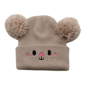 Detail of beige knitted baby beanie with dual pom-poms and cute cartoon face embroidery