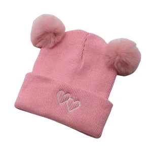 Close-up of pink knit baby hat with double pompoms and white heart embroidery detail