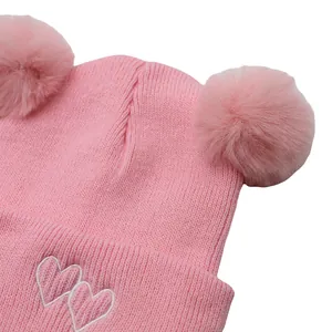 Close-up of pink knitted hat featuring white heart embroidery and dual faux fur pom-poms