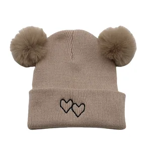 Close-up of beige knit hat featuring double fluffy pom poms and heart embroidery