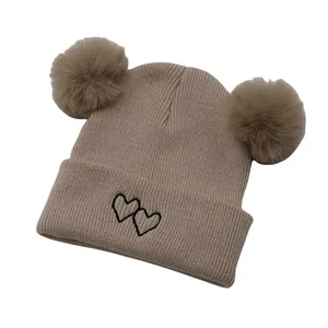 Detail of khaki knit beanie with dual pom-poms and black heart embroidery on the cuff
