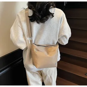 Detail view of a beige nylon crossbody bag worn over a gray sweatshirt in a minimal setting