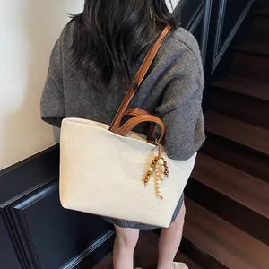 Large beige canvas tote bag with brown leather handles worn by a model in a stairwell