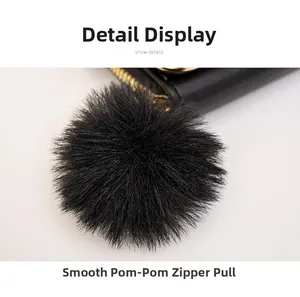 Detail of smooth gold zipper with large black fluffy pom-pom on leather crossbody bag