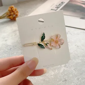 Hand holding a pink flower hair clip with green leaves on a white card display