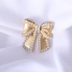 Matte gold bow brooch with pearl edges on white fabric, elegant fashion accessory
