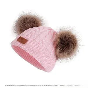 Détail d'un bonnet en maille rose pour bébé avec deux pompons en fourrure et écusson cuir
