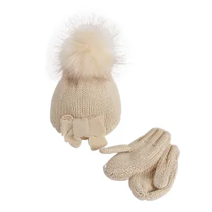 Detailed view of beige baby knit hat with fluffy pompom, bow, and matching mittens