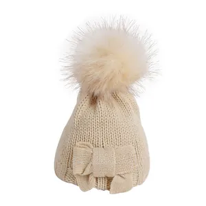 Detailed knit texture of a beige baby beanie with a faux fur pompom and bow accent