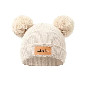 Detail van de beige gebreide babymuts met mini lederen patch en twee zachte pompons