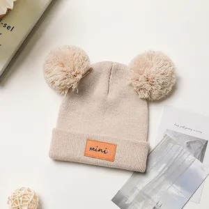 Crèmekleurige gebreide babymuts met dubbele pompons en leren mini-patch