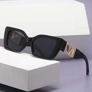 Square black sunglasses with gold V-shaped rhinestone temples on a white geometric stand