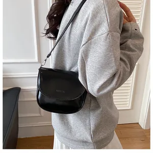 Glossy black saddle bag with embossed logo worn by a model in casual grey and white attire