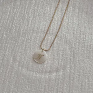 Close-up of golden snake chain necklace with round white shell pendant and letter H detail