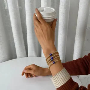 Close-up of four stacked gold beaded evil eye bracelets worn by a model holding a cup