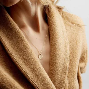 Macro detail of hammered gold coin pendant on a delicate chain worn over brown fabric