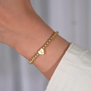 Macro detail of gold-plated beaded bracelet featuring a heart charm with engraved letter X