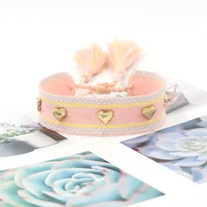 Dettaglio di bracciale intrecciato rosa con borchie a cuore dorate e nappe boho