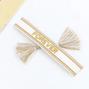 Detail of hand-woven white bracelet with golden Forever embroidery and bohemian tassels
