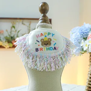 Detail of plush cream pet birthday bandana with embroidered bear and rainbow tassels