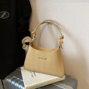 Detail of beige woven shoulder bag with knotted straps resting on a book and silver suitcase