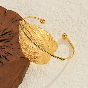 Goldener Armreif mit hohlem Blattdesign und grünen Edelsteinen auf Holzuntergrund