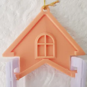 Close-up of peach house-shaped plastic ornament with gold string on white brick wall