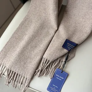 Close-up of oatmeal wool scarf with fringe edge and brand tag on leather surface